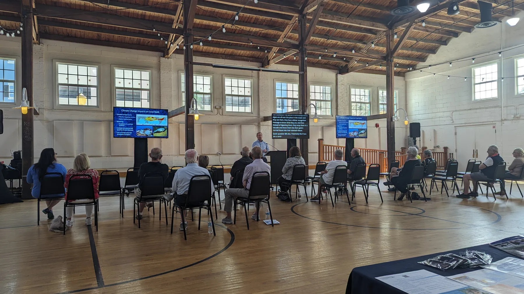 OLLI at FAU lecture on climate change and coral reefs in the Field House at Old School Square, with presentation slides on display screens on both sides of the speaker