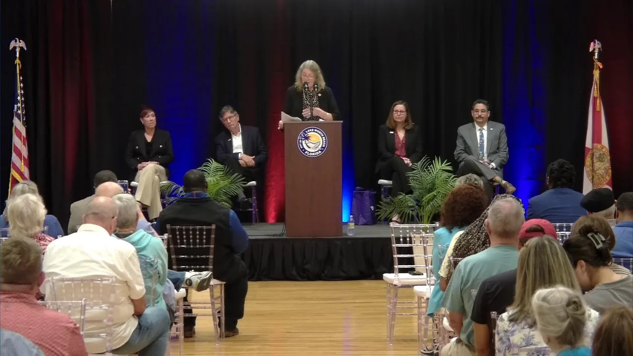 Speaker at the podium in front of pipe and drape, with city officials seated on stage and a full audience, at the Lake Worth Beach 2025 State of the City